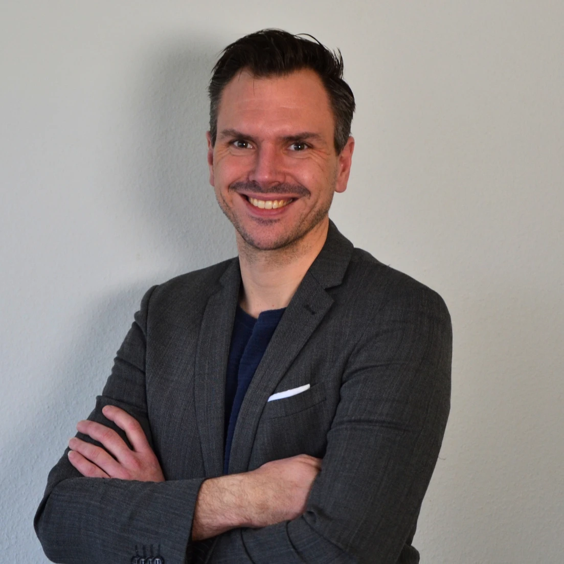 Ein Mann in einem Anzug mit verschränkten Armen lächelt.