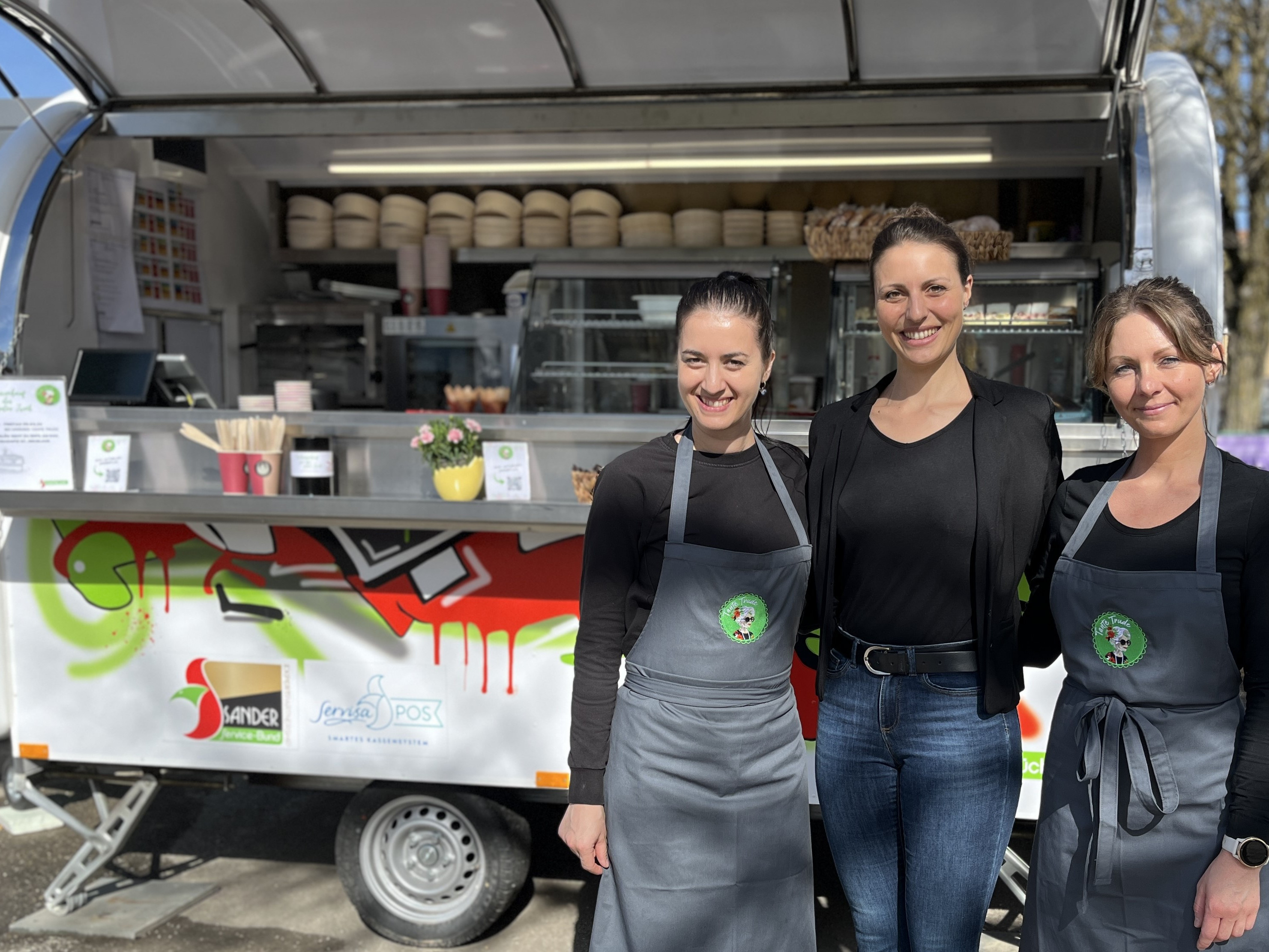 Drei Frauen stehen vor einem zum Foodtruck umfunktionierten Anhänger und lächeln.